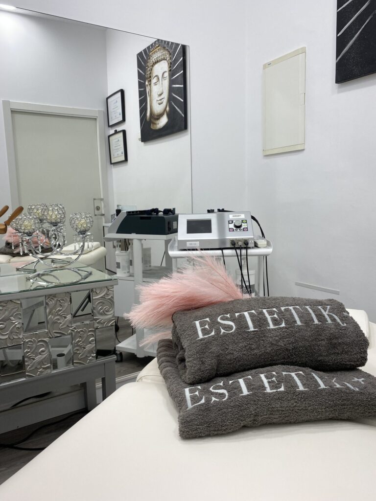 Camilla blanca con toallas grises ESTETIK+ apiladas, espejo grande, equipo estético en carro y cuadro de Buda.