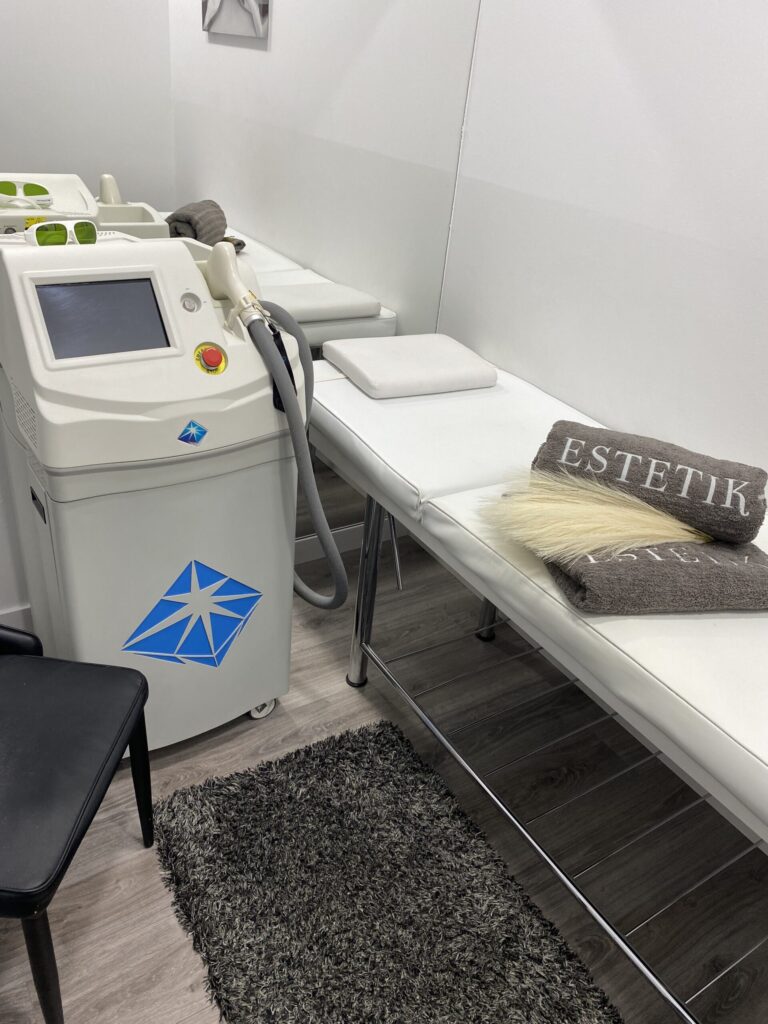 Equipo de depilación láser junto a camillas blancas, toalla ESTETIK+ y alfombra gris en una cabina de paredes blancas.