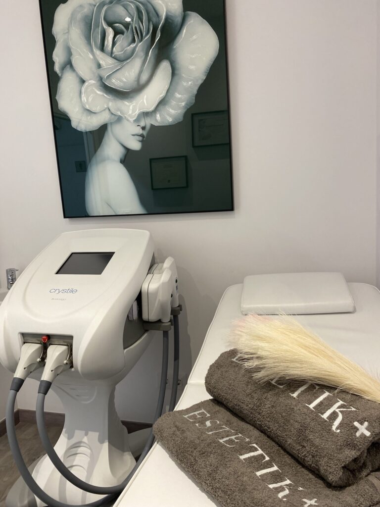 Camilla blanca con toallas grises ESTETIK+, equipo estético con pantalla y gran cuadro de mujer con flor en la pared.