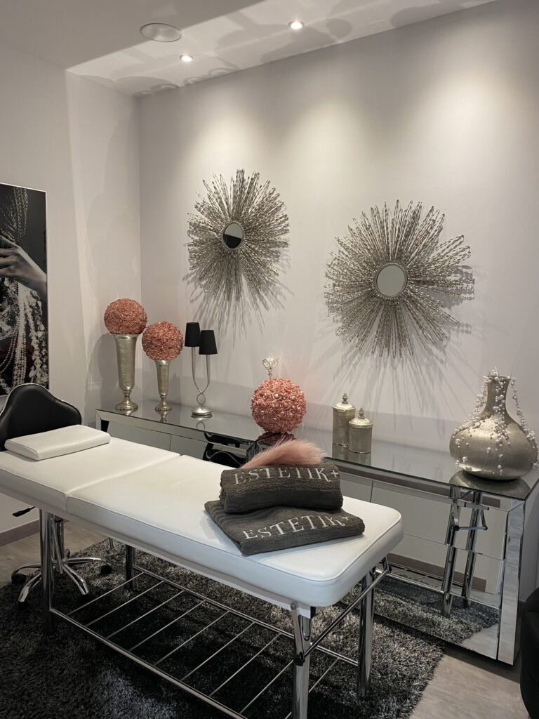 Camilla blanca con toallas ESTETIK+ en cabina; tocador espejado y dos espejos sol sobre pared clara.