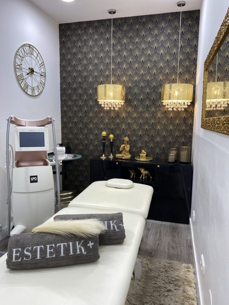 Camilla blanca con toallas ESTETIK+, equipo LPG y lámparas doradas en cabina estética.