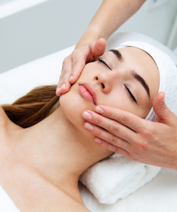 Mujer recostada con toalla bajo la cabeza recibe masaje facial con ambas manos en una cabina de estética.