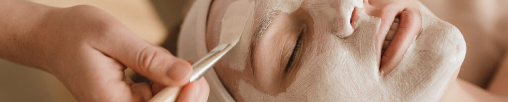 Mujer recibiendo una mascarilla facial aplicada con brocha mientras está recostada en una camilla de estética.
